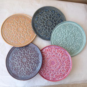 Può includere: Un set di cinque sottobicchieri circolari in ceramica, ognuno caratterizzato da un intricato motivo a mandala in rilievo. I sottobicchieri sono rifiniti in colori distinti: giallo senape, grigio antracite, verde acqua chiaro, viola scuro e rosa.