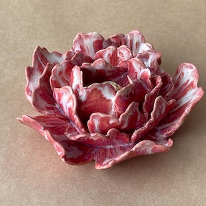 May include: A ceramic flower sculpture featuring layered, ruffled petals in a marbled blend of deep red, pink, and white. The glossy finish highlights the intricate, organic curves of the bloom, which is shaped to resemble a full, open peony.