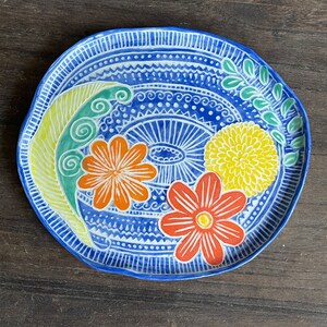 May include: A blue ceramic plate with a white and blue patterned design. The plate features three flowers in orange, yellow, and red, with green leaves.