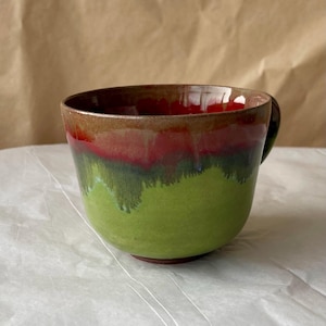 May include: A ceramic mug featuring a wide, rounded bowl shape with a handle. The exterior glaze transitions from a vibrant lime green at the base to a deep, saturated red in the middle, topped with a smooth, earthy brown rim at the top edge.