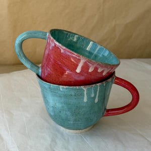 May include: Two stacked ceramic mugs featuring a contrasting colour scheme. The top mug is red with a teal interior and handle, while the bottom mug is teal with a red handle. Both pieces exhibit a speckled texture and white drip glaze details.