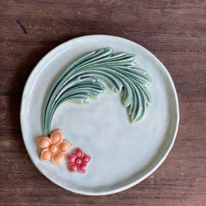 May include: A light blue ceramic plate with a raised green leaf design and three raised flowers in orange, red, and yellow.