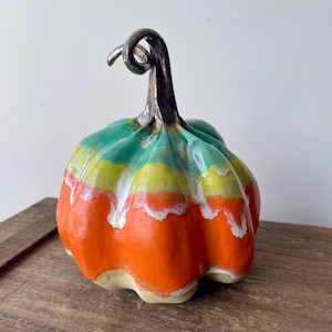 May include: A ceramic pumpkin in a bright colour scheme. The pumpkin has horizontal stripes of orange, yellow, and green, with white accents. The stem is a dark, metallic colour. A decorative autumn-themed item.