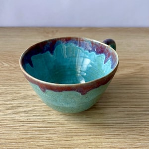 May include: A handmade ceramic bowl with a turquoise interior and a reddish-brown rim. The bowl has a small handle and a speckled, light green exterior. The bowl is sitting on a wooden surface.