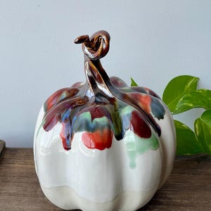 May include: A ceramic pumpkin with a white body and a stem with brown and red glaze. The top of the pumpkin has streaks of red, green, and blue glaze. The pumpkin is a decorative item.