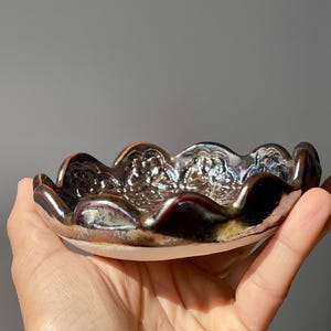 May include: A small, iridescent ceramic dish with a scalloped rim. The dish has a dark, metallic finish with hints of brown and silver. The interior has a textured, decorative pattern. The base of the dish is a light, creamy colour.