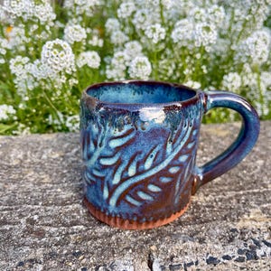 Puede incluir: Una taza de cerámica hecha a mano con un esmalte azul y marrón. La taza presenta un diseño de hojas y un asa curvada. La taza está sobre una superficie de hormigón con flores blancas en el fondo.