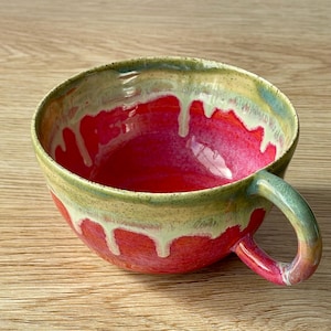 May include: A handmade ceramic bowl with a handle. The interior is a vibrant red, with a green and yellow rim. White glaze drips down the sides, creating a unique, artistic look. The bowl is perfect for serving soups or stews.