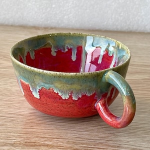 May include: A handmade ceramic mug with a vibrant red interior and a handle. The rim features a green and blue glaze that drips down the sides, creating a unique, artistic effect. The mug is designed for hot beverages.