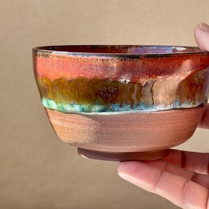 May include: A small ceramic bowl with a rounded shape. The upper portion features a glossy, multi-coloured glaze in shades of vibrant red, earthy brown, and teal blue. The lower portion of the bowl is made of unglazed, textured terracotta clay.