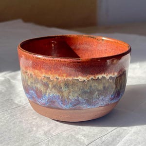 May include: A small ceramic bowl with a smooth, unglazed terracotta base and a glossy, multi-coloured glaze. The upper portion features a vibrant gradient of deep red, transitioning into earthy brown, olive green, and hints of blue at the rim.