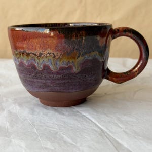 May include: A ceramic mug featuring a wide, rounded body and a curved handle. The upper portion displays a glossy glaze with blended hues of deep red, dark brown, and hints of purple, while the base is a matte, unglazed dark brown clay.
