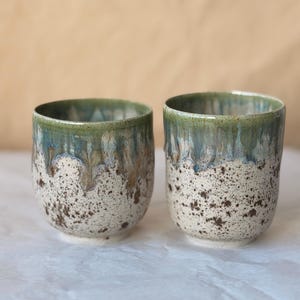 May include: Two ceramic cups featuring a speckled cream-coloured base with dark brown flecks. The upper portion of each cup is finished with a reactive glaze that transitions from a deep blue to a muted green, creating a dripping effect.