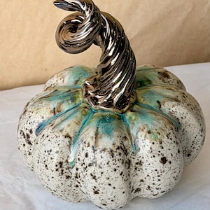 May include: A ceramic pumpkin sculpture featuring a speckled cream and white body with teal blue glaze accents dripping from the top. The pumpkin is topped with a prominent, spiraled, metallic dark bronze stem with a glossy, reflective finish.