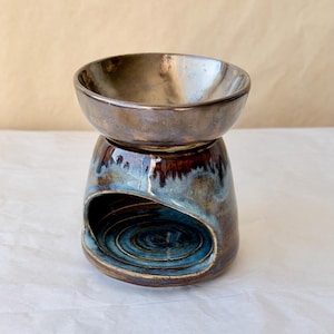 May include: A ceramic wax warmer featuring a glossy metallic brown bowl atop a base with a wide arched opening. The base displays a reactive glaze with swirling patterns of deep blue, brown, and cream, while the interior floor shows concentric rings.