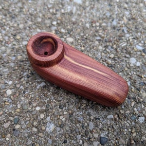 Puede incluir: Una pipa de madera artesanal de color marrón rojizo con un cuenco redondeado. La pipa presenta un acabado liso y pulido y una forma cilíndrica, mostrando la veta natural de la madera.