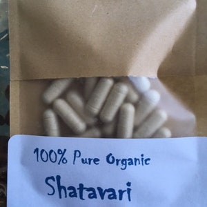 Puede incluir: Una bolsa de papel blanca con una etiqueta azul que dice "100% Pure Organic Shatavari". La bolsa contiene un gran n&uacute;mero de c&aacute;psulas blancas.