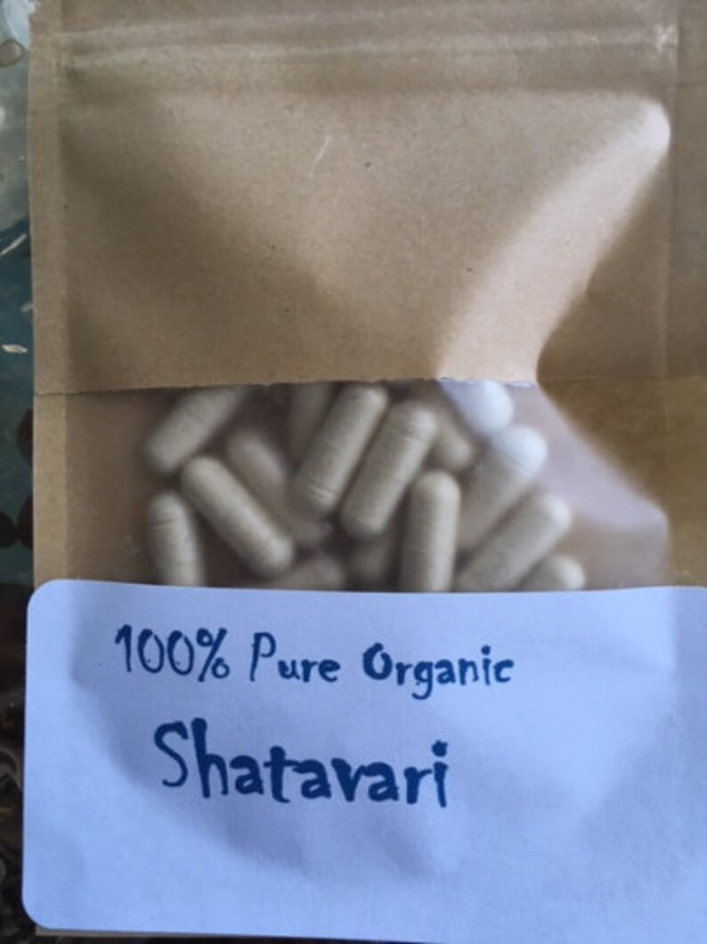 Puede incluir: Una bolsa de papel blanca con una etiqueta azul que dice "100% Pure Organic Shatavari". La bolsa contiene un gran n&uacute;mero de c&aacute;psulas blancas.