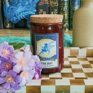 May include: A red glass candle jar with a cork lid and a blue label that says "Fiction & Flames Pixie Dust" with a fairy illustration. The candle is on a chequered tray with a bouquet of purple flowers.