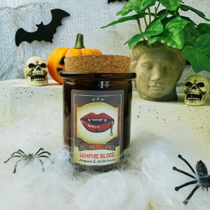 May include: A brown glass candle jar with a cork lid. The candle jar has a label that says "Fiction & Flames Vampire Blood Pomegranate & Red Velvet Vanilla". The label features a red vampire mouth with fangs and dripping blood. The candle jar is sitting on a white fluffy surface with a black spider and a small pumpkin.