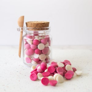 May include: A glass jar filled with pink and white chocolate chips, with a wooden spoon resting on the rim. The jar has a cork lid and is sitting on a white surface with a speckled pattern. Some chocolate chips have spilled out of the jar.