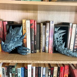 May include: A pair of blue dragon bookends on a bookshelf with a variety of books. The bookends are made of a textured material and have a realistic dragon design.
