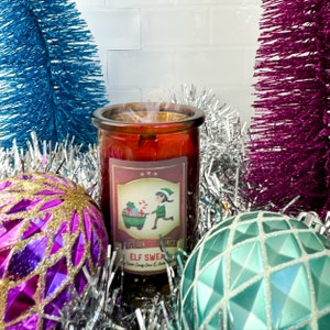 May include: A red glass candle jar with a white label that says "Elf Sweets" and features a cartoon elf pushing a wheelbarrow full of presents. The candle is surrounded by silver and purple tinsel.