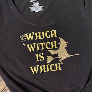 Op de afbeelding: Zwart T-shirt met gouden glitter-lettering die "Which Witch is Which" zegt en een silhouet van een heks die op een bezem vliegt.