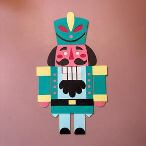 Op de afbeelding: Een platte, tweedimensionale notenkraker-uitsparing gemaakt van gelaagd papier of karton. De figuur heeft een groenblauwe hoed met een gele pluim, een lichtblauw jasje, een zwarte snor en baard, witte tanden en roze accenten op de wangen.
