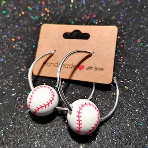 May include: Silver hoop earrings with a white baseball charm with red stitching. The earrings are on a brown card with the text "Handmade with love".