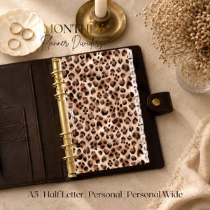 May include: A dark brown leather ring binder planner featuring monthly dividers with a leopard print pattern. The gold-toned metal rings hold the pages, and the side tabs are labeled with the months of the year from January to December.