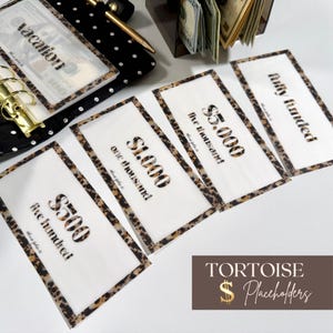 May include: A set of rectangular translucent cash envelopes with a leopard print border. The envelopes feature black text reading $500 five hundred, $1,000 one thousand, $5,000 five thousand, and fully funded. A black polka dot binder and gold pen are visible.