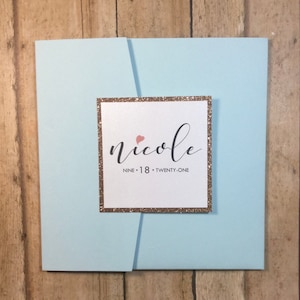 May include: A light blue invitation card with a gold glitter border. The card has a white insert with the name "nicole" in black cursive writing and a small pink heart. The text "NINE - 18 - TWENTY-ONE" is printed below the name.