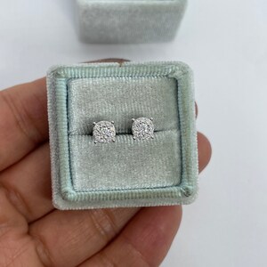 May include: A pair of silver stud earrings with a round, pave-set diamond design. The earrings are in a small, light blue velvet box.
