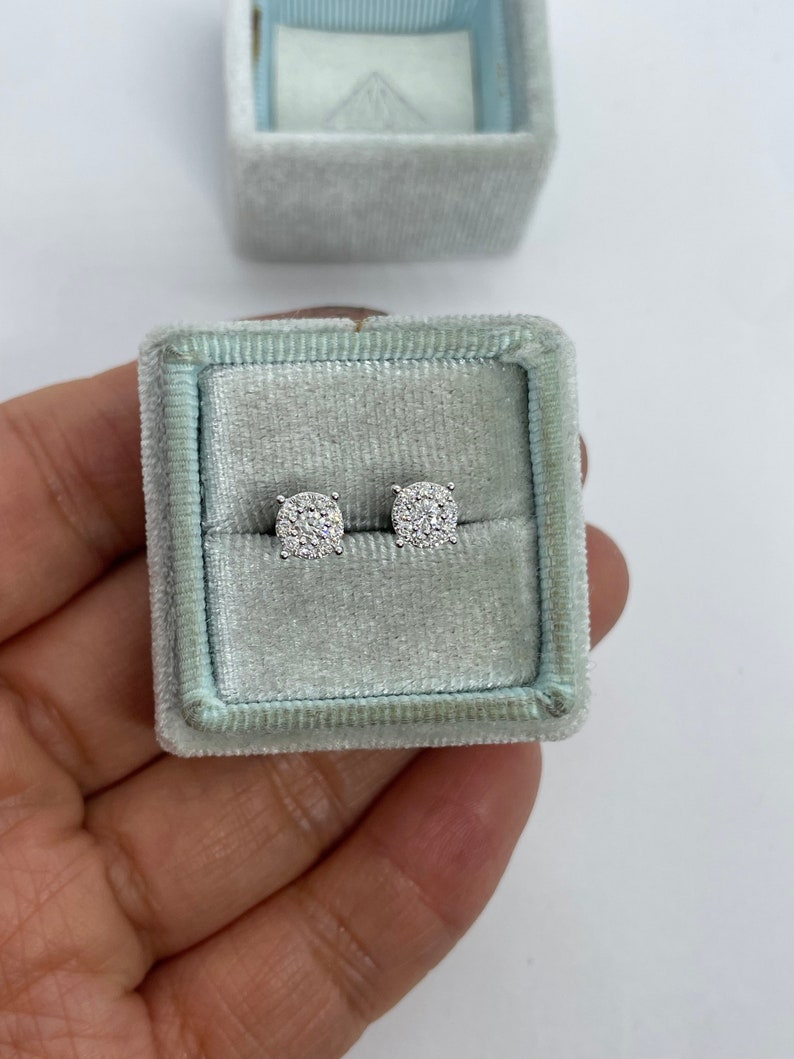May include: A pair of silver stud earrings with a round, pave-set diamond design. The earrings are in a small, light blue velvet box.