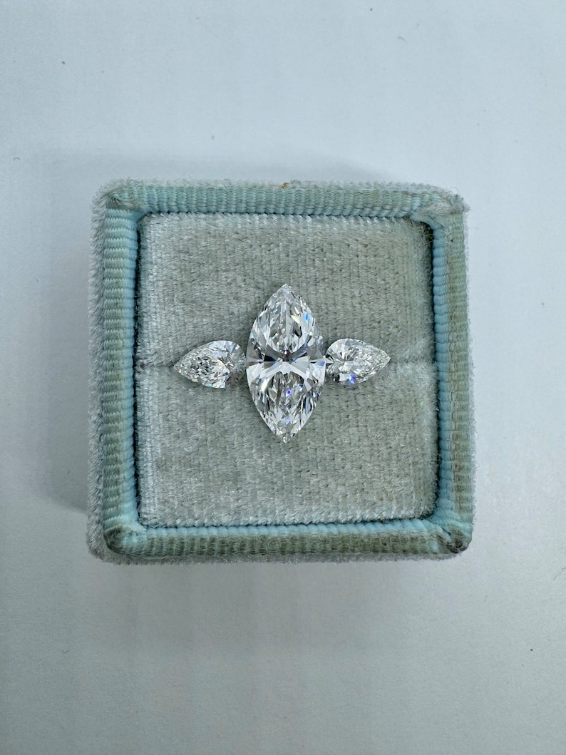 May include: A three-stone engagement ring with a large marquise-shaped center stone and two smaller pear-shaped side stones. The ring is set in a white metal band and is displayed in a blue velvet ring box.