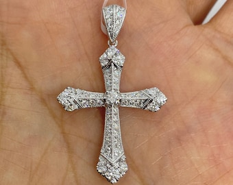 May include: A silver pendant in the shape of a cross, encrusted with numerous small, sparkling gemstones. The cross has a detailed, ornate design with a bail at the top for a chain. The pendant is held up by a white string.