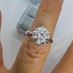 May include: A round-cut diamond ring with a silver band. The large, clear diamond is held in place by six prongs. The ring is shown on a finger, with a light blue ring box in the background.