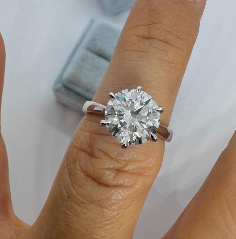 May include: A round-cut diamond ring with a silver band. The large, clear diamond is held in place by six prongs. The ring is shown on a finger, with a light blue ring box in the background.