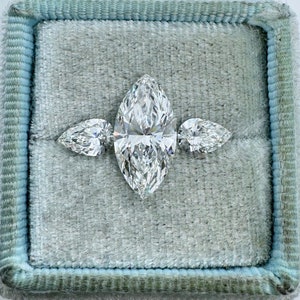 May include: A three-stone engagement ring with a large marquise-shaped diamond in the center and two pear-shaped diamonds on either side. The ring is displayed in a light blue velvet ring box.