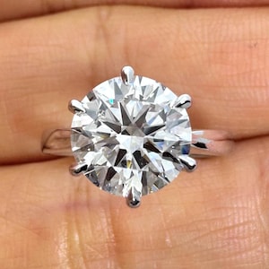 May include: A close-up of a sparkling, round-cut diamond ring. The large, clear gemstone is set in a silver band with six prongs. The ring is held in a hand, showcasing its brilliance and design.