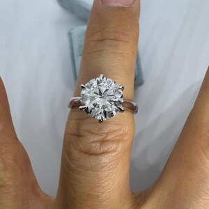 May include: A silver ring with a large, round, clear gemstone. The ring is set with six prongs, and the gemstone sparkles brightly. The ring is on a finger, with a light blue ring box in the background.