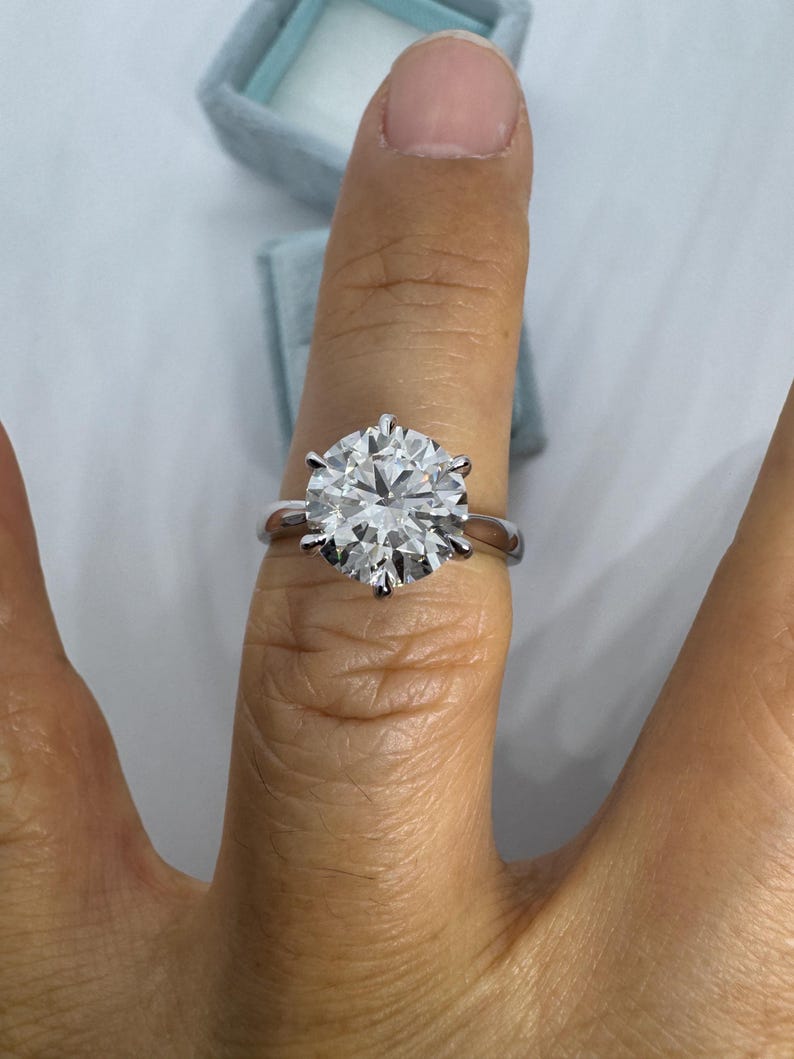 May include: A silver ring with a large, round, clear gemstone. The ring is set with six prongs, and the gemstone sparkles brightly. The ring is on a finger, with a light blue ring box in the background.