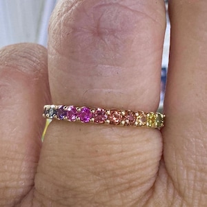 May include: A gold band ring featuring a row of colorful gemstones in a rainbow spectrum. The gemstones transition from purple to pink, orange, and yellow. The ring is shown on a finger.