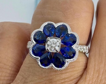 May include: A floral-shaped ring featuring a central square-cut diamond surrounded by pear-shaped blue sapphires. The ring has a white metal band with small accent stones, creating a striking contrast of colors and textures. The design is elegant and eye-catching.