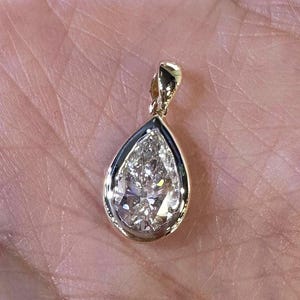 May include: A pear-shaped diamond pendant with a gold bail and a dark border. The diamond is clear and faceted, reflecting light. The pendant is likely a piece of jewelry.