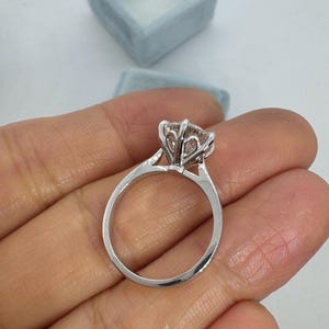 May include: A silver engagement ring with a large, clear gemstone held in a six-prong setting. The ring is held in a hand, with a light blue ring box in the background.