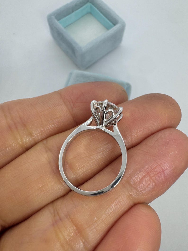 May include: A silver engagement ring with a large, clear gemstone held in a six-prong setting. The ring is held in a hand, with a light blue ring box in the background.