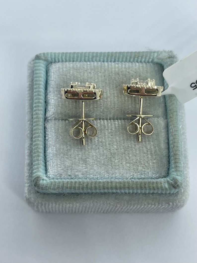 May include: A pair of gold earrings with a square-shaped diamond in the center surrounded by smaller diamonds. The earrings are in a blue velvet box.
