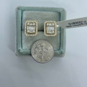 May include: A pair of gold earrings with a square shape and a row of baguette diamonds in the center. The earrings are in a blue velvet box with a dime for scale. The box has a label that reads "ET5P01A5S P 320615".